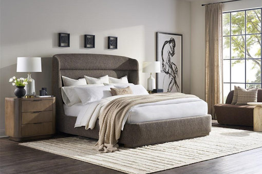 Modern bedroom with a large upholstered bed, neutral bedding, two wooden nightstands with lamps, wall art, a rug, and a window with beige curtains letting in natural light.