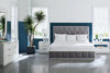 Modern bedroom with a gray upholstered bed, white bedding, two nightstands with lamps, a dresser, and a tall indoor plant. Large windows allow natural light into the space.