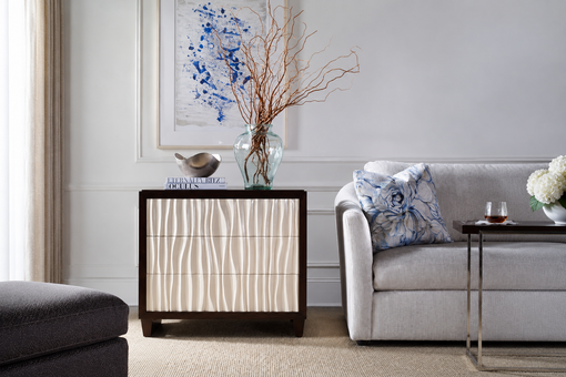Luxury modern accent cabinet with textured white wave-pattern doors and a dark wood frame, styled with decorative books, a glass vase of branches, and metallic accents beside a gray sofa in a bright contemporary living room.