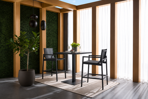 Modern dining area with two black high chairs and table on a beige rug. Sunlight filters through sheer curtains and greenery adds a fresh touch.