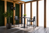 Modern dining area with two black high chairs and table on a beige rug. Sunlight filters through sheer curtains and greenery adds a fresh touch.