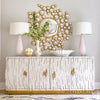 A white textured sideboard with gold handles, two pink lamps, a vase with flowers, a scalloped bowl, and a decorative round mirror with gold accents on a light wall.