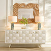 Modern white sideboard with geometric drawer fronts, paired with blue ceramic table lamps, a woven rattan wall mirror, and decorative greenery in a bright luxury living room.