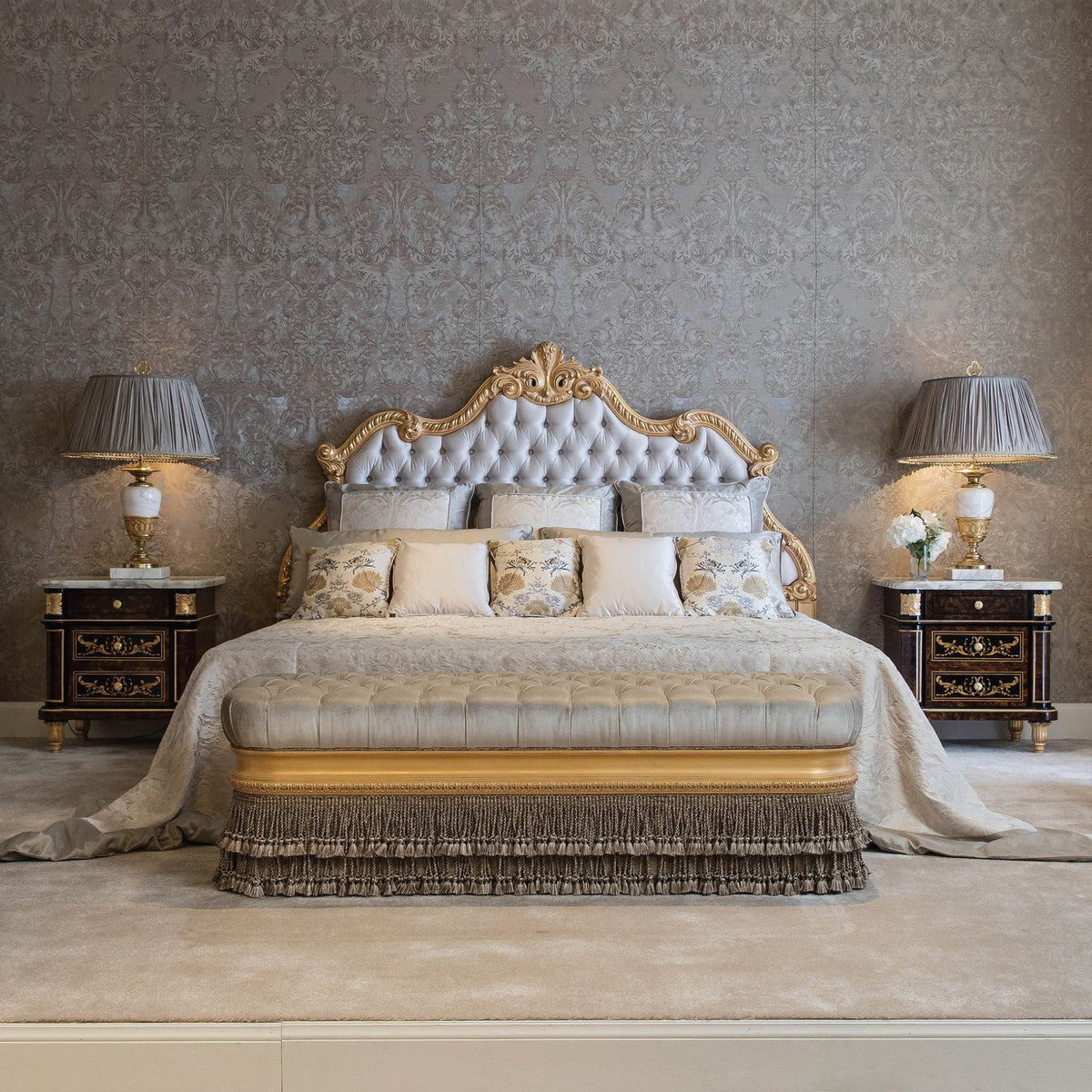 A luxurious bedroom scene featuring a Classico Bed with a padded headboard, upholstered in a beige fabric, flanked by two nightstands with gold-colored accents, on a carpeted floor with a gold-colored cushion on the bed.